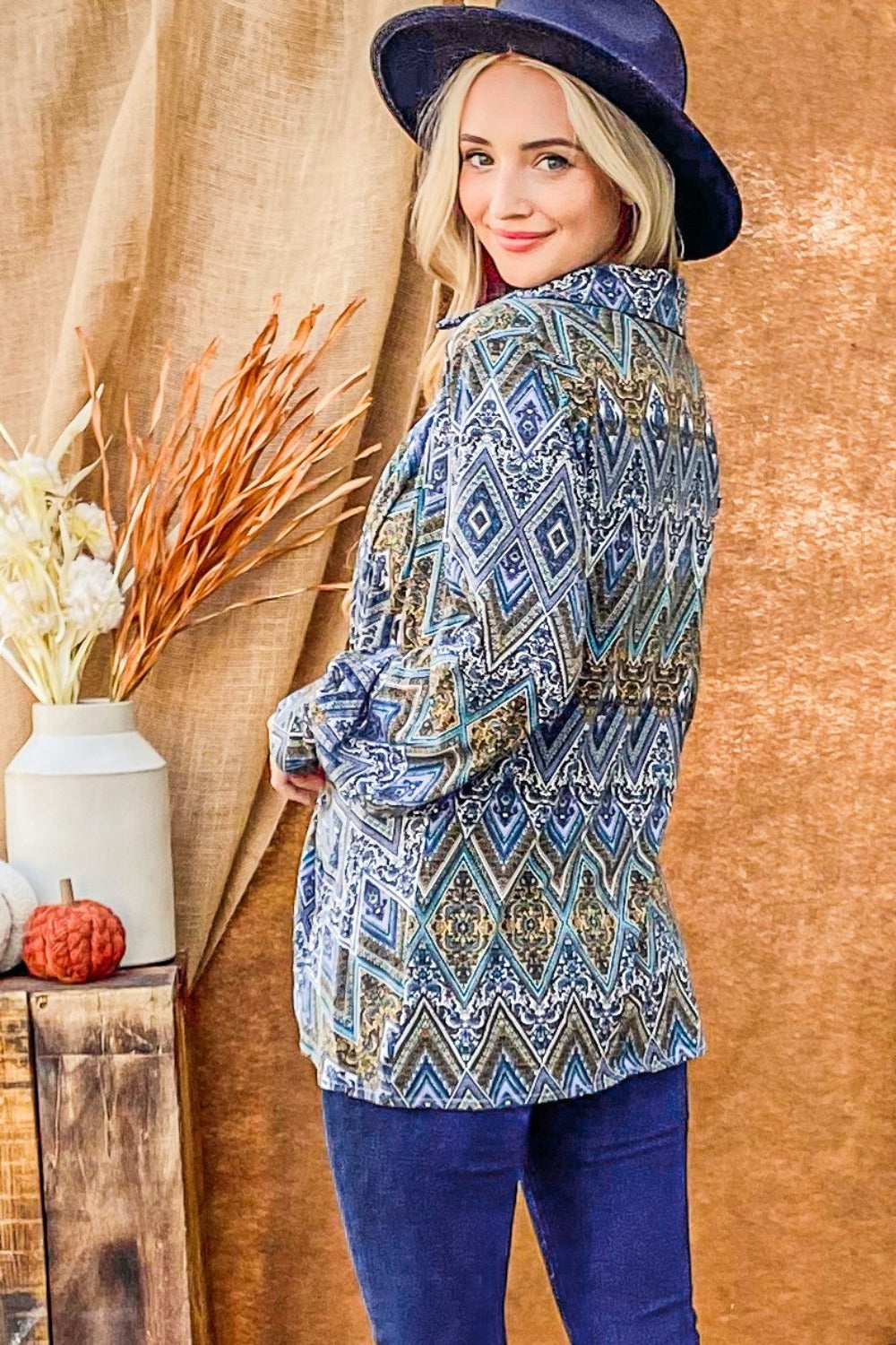 A woman with long blonde hair wearing an And The Why Print Twist Knot Long Sleeve Blouse, blue jeans, and a dark blue hat stands in front of a brown backdrop.