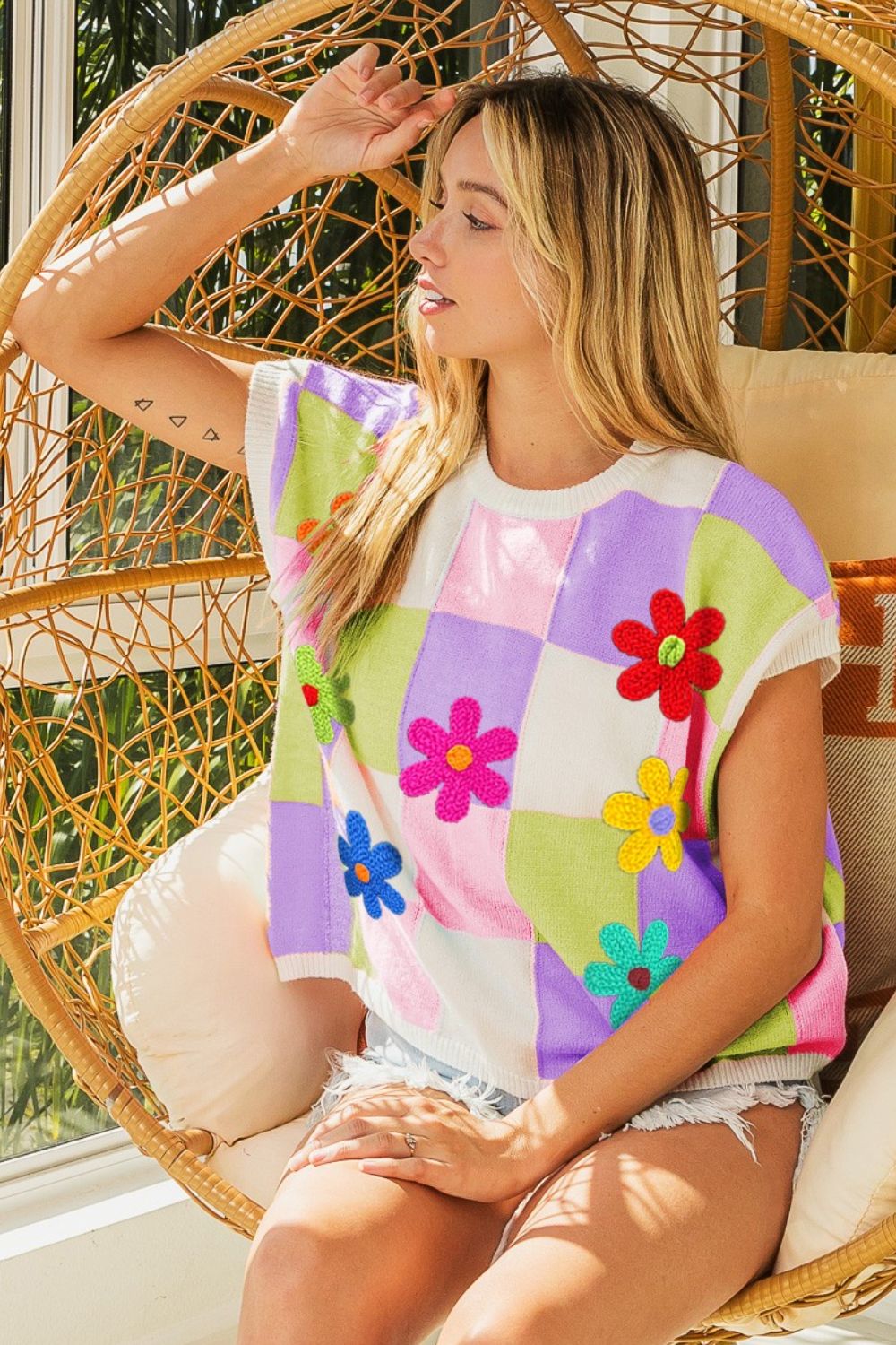 A woman sits in a rattan hanging chair, wearing a BiBi Flower Patch Checkered Sweater Vest with denim shorts. She has long blonde hair and is looking to the side, with her hand resting on her head, epitomizing a cozy layered ensemble perfect for a trendy fall wardrobe.