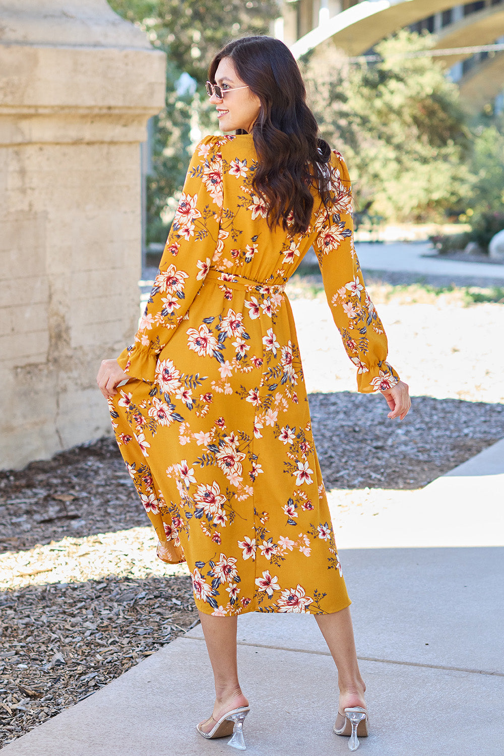 A woman wearing the Double Take Full Size Floral Tie Back Flounce Sleeve Dress in yellow, along with sunglasses, is walking on a paved path outdoors. She is smiling and has one hand on her hip.