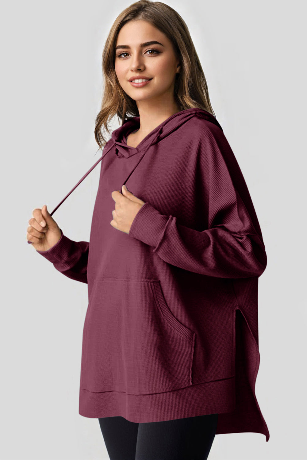 A woman in a Red Dahlia Waffle Knit Fleece Lined High Low Oversized Hoodie and black shorts is seen from behind, standing against a plain gray background.