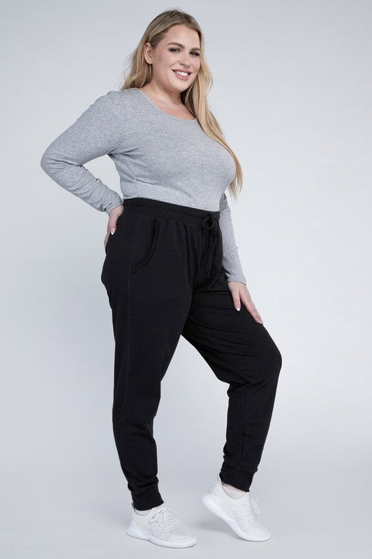 A person stands against a white background, wearing a white t-shirt, grey Plus-Size Jogger Pants with an adjustable waistband, and white sneakers. They have long blonde hair and are posing with one hand on their hip.