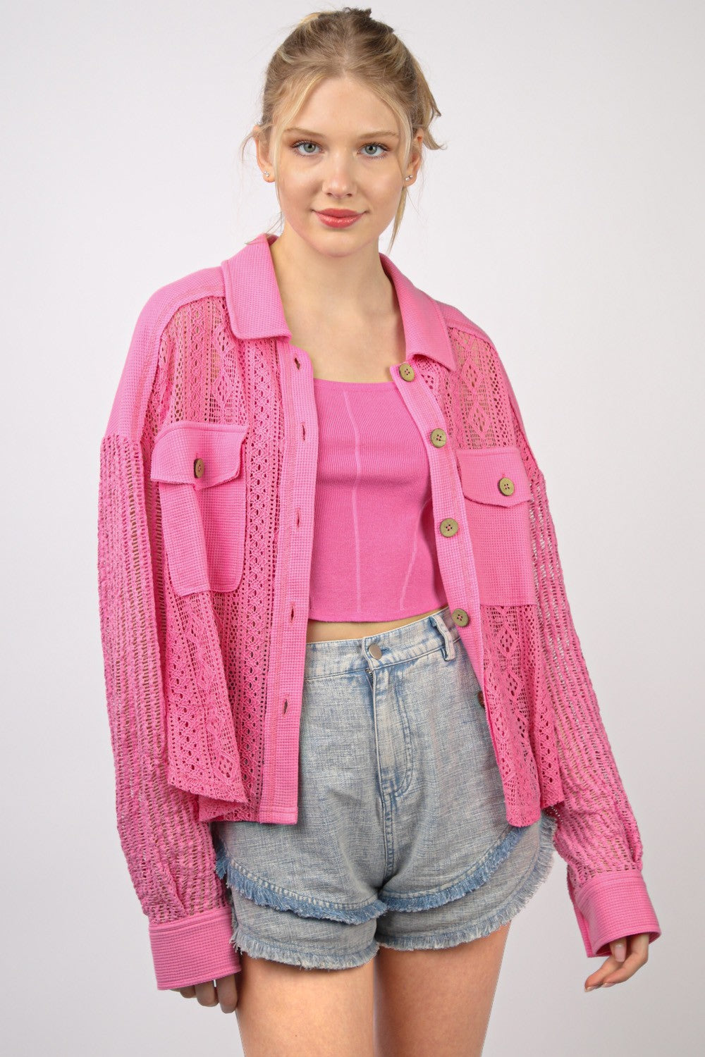 A person wearing a bright pink, long sleeve button-up lace shirt with drop shoulders and high-waisted denim shorts stands against a plain background.