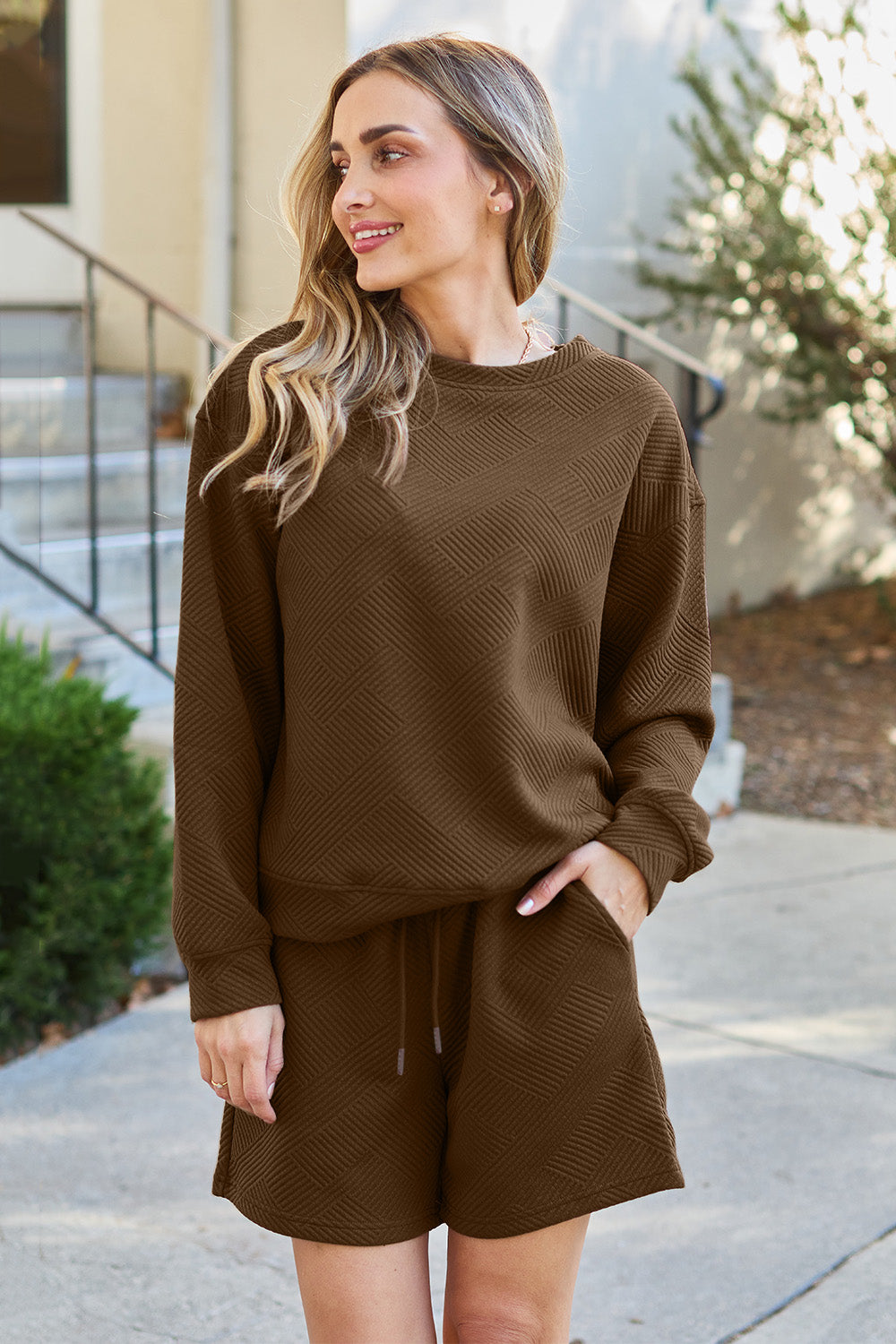 A woman decked out in the Double Take Full Size Texture Long Sleeve Top and Drawstring Shorts Set, featuring a brown textured two-piece with a long-sleeve top and matching pocketed shorts, smiles at the camera.