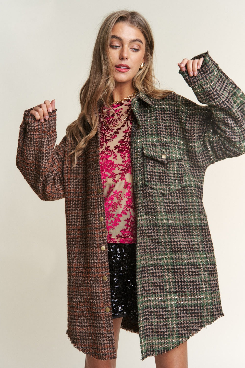 A woman wearing an eclectic mix poses against a plain background, showcasing an ideal fall wardrobe ensemble. She dons a mixed-patterned oversized jacket over a red floral sheer top and black sequined shorts. The J.NNA Snap Down Tweed Plaid Contrast Longline Shacket adds texture and depth to her look.