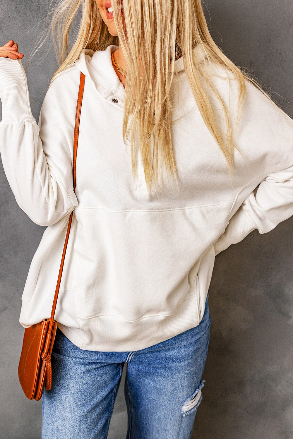 A person with long blonde hair, wearing a White Batwing Sleeve Pocketed Henley Hoodie and blue jeans, stands with their back to the camera, holding a small brown bag over their shoulder. The hoodie features a subtle patchwork design that adds an interesting texture to the outfit.