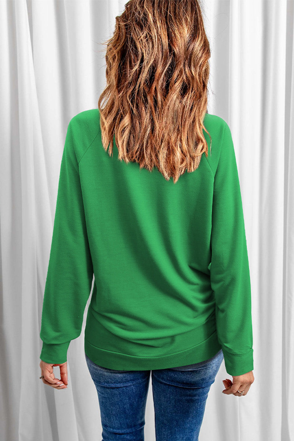 A person with wavy hair wearing a Green Solid Round Neck Raglan Sleeve Sweatshirt and blue jeans is standing with their back to the camera in front of white curtains.