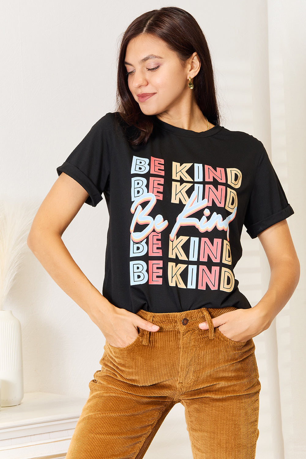 A woman stands with her hands in the pockets of her brown corduroy pants, wearing a casual Simply Love BE KIND Graphic Round Neck T-Shirt that reads "BE KIND" in large colorful letters, showcasing positive message clothing.