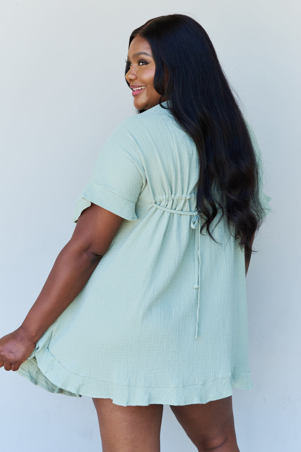 A woman is wearing the Ninexis Out Of Time Full Size Ruffle Hem Dress with Drawstring Waistband in Light Sage. Her long, dark hair falls gracefully as she smiles and looks to her left, standing against a plain background.