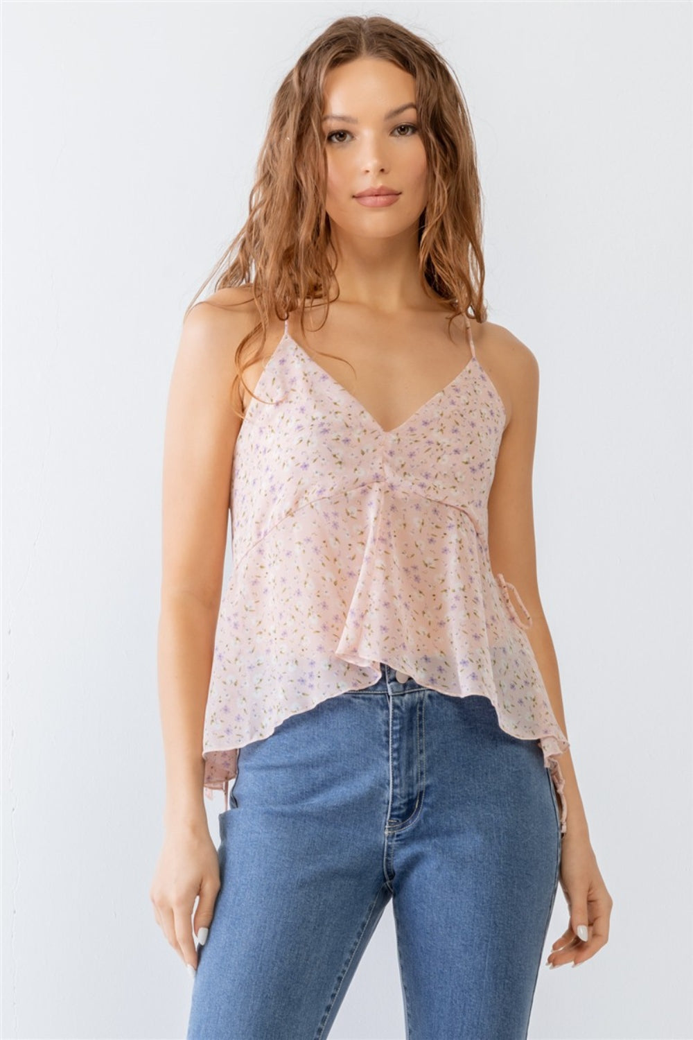 A woman wearing the Miss Love / Kevin Co. Floral Print V-Neck Criss-Cross Back Flare Cami and blue jeans stands against a plain white background.