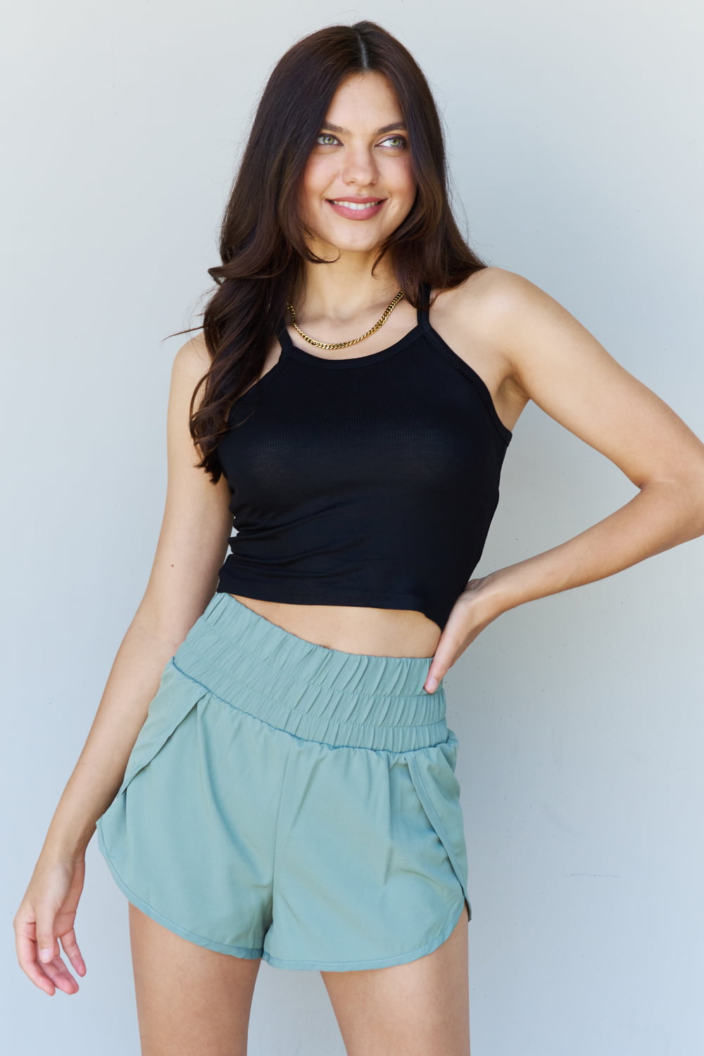 A person wearing a Ninexis Everyday Staple Soft Modal Short Strap Ribbed Tank Top in Black and light blue shorts is standing against a plain background, smiling slightly and looking to the side.