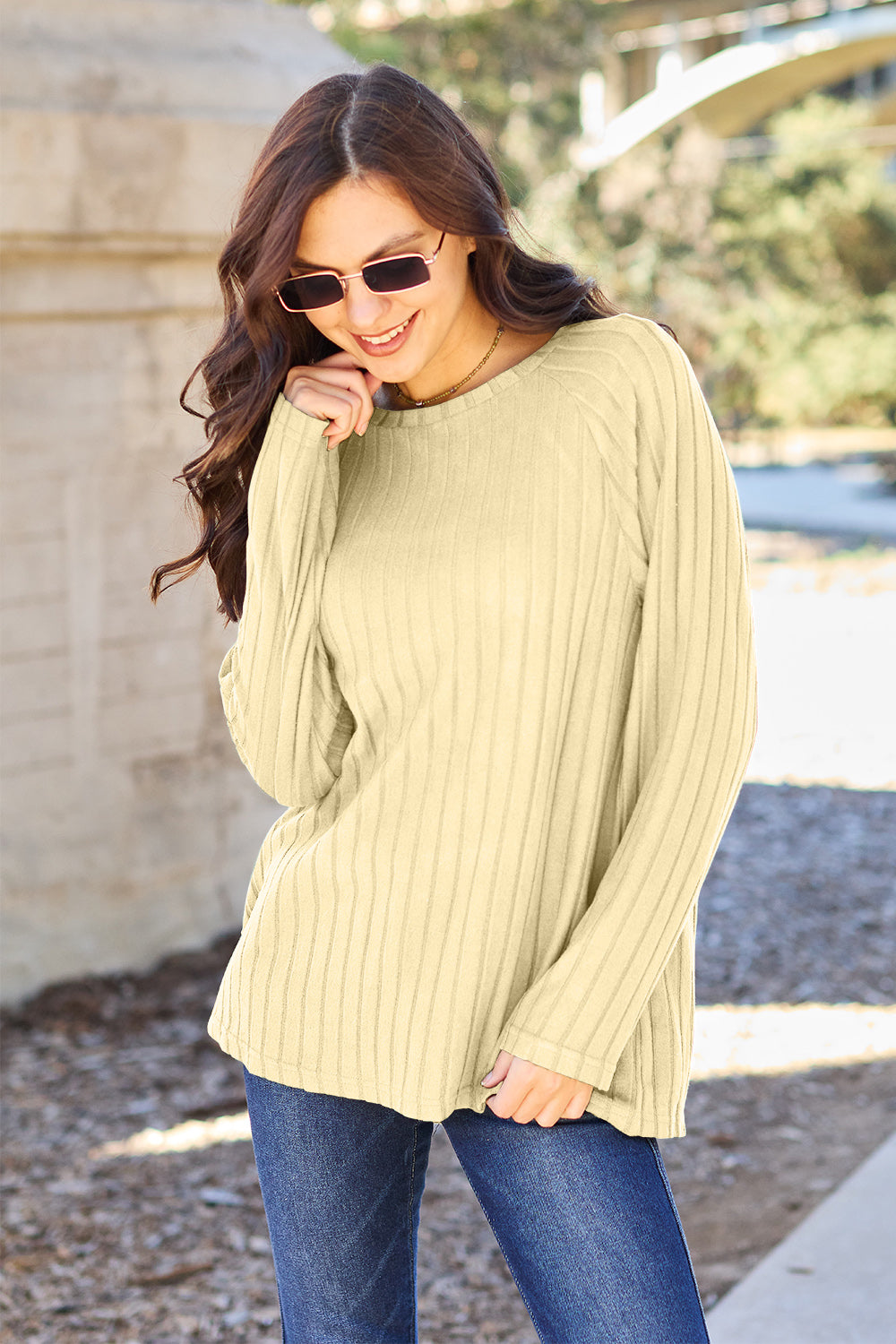 A woman with long brown hair wearing sunglasses and a Basic Bae full-size, slightly stretchy ribbed round neck long sleeve knit top in red, paired with blue jeans, is standing outdoors.