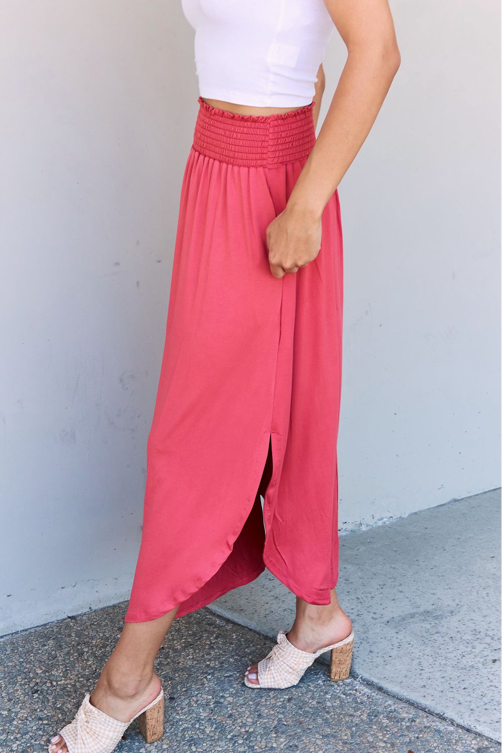 A person wearing a white top and the Doublju Comfort Princess Full Size High Waist Scoop Hem Maxi Skirt in an elegant coral color, paired with tan sandals, stands in front of a plain wall.