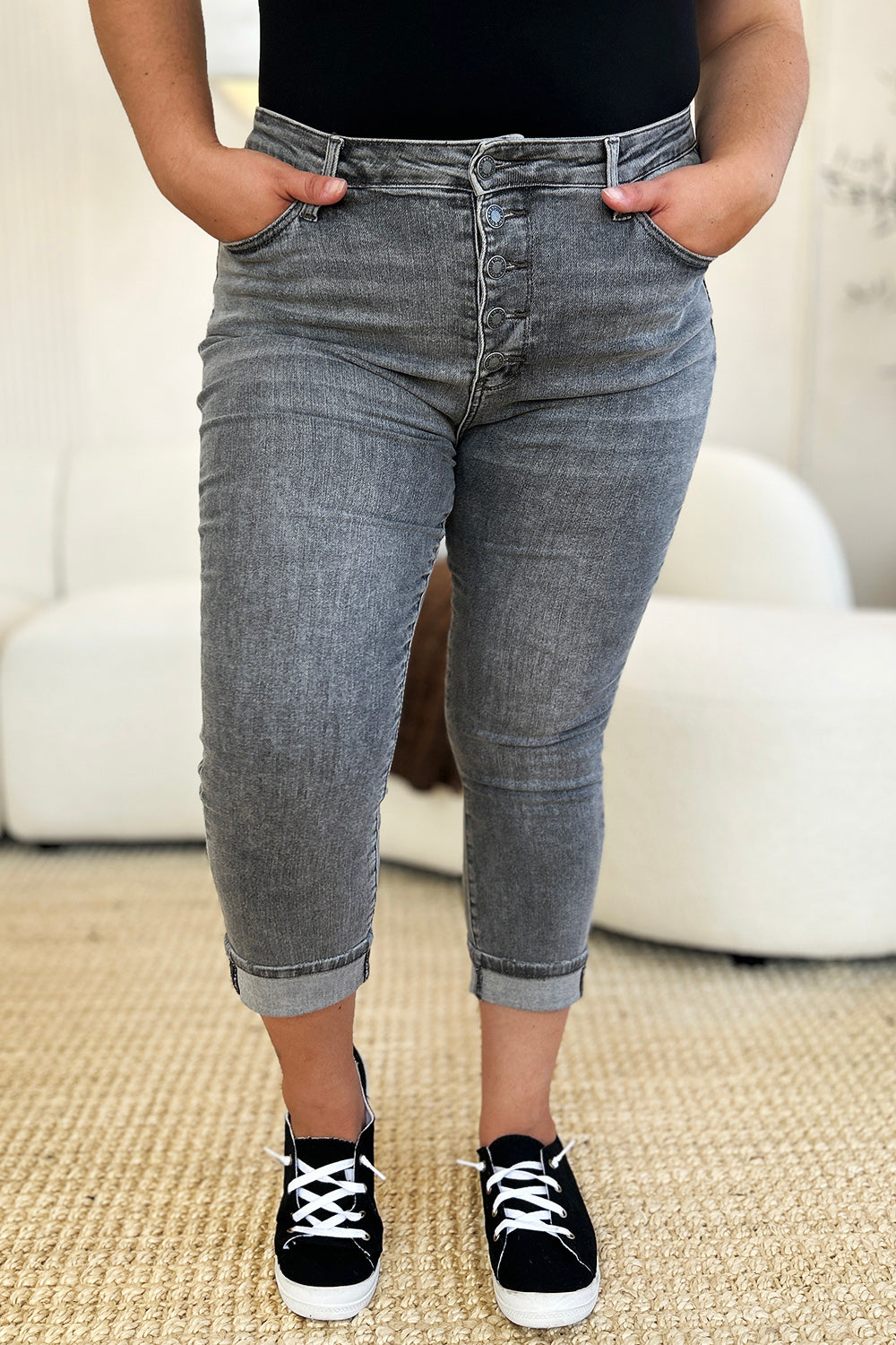 A person wearing Judy Blue Full Size Button Fly High Waist Cuffed Capris in gray, paired with a white top and white sneakers, stands on a beige carpeted floor.