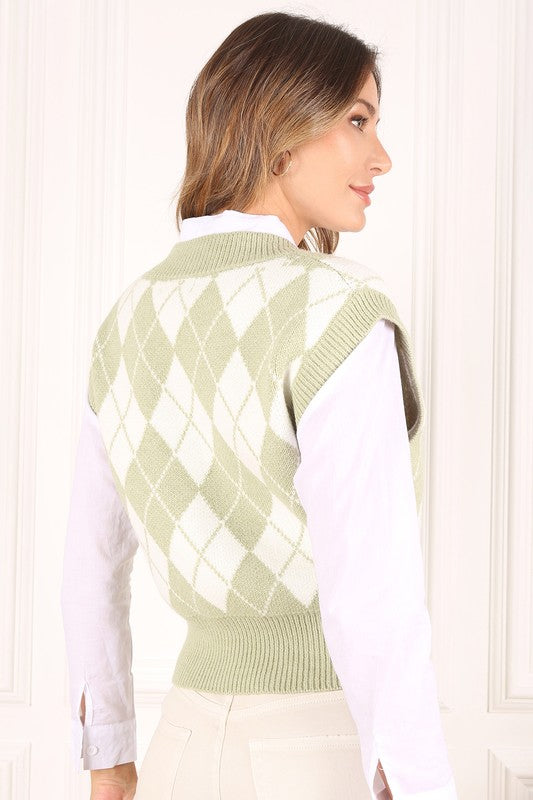 A person wearing a knitted argyle sweater vest in light green over a white shirt stands against a neutral background.