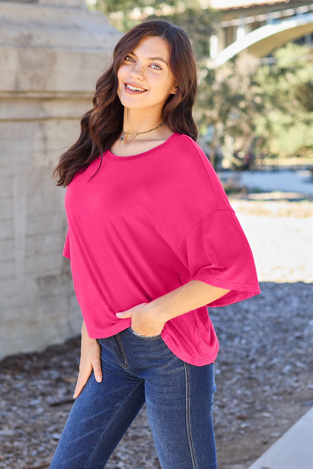 A person with long dark hair and a bright smile stands outdoors, showcasing their basic style in a loose, slightly stretchy *Basic Bae Full Size Round Neck Drop Shoulder T-Shirt* in pink and blue jeans, with one hand on their hip.