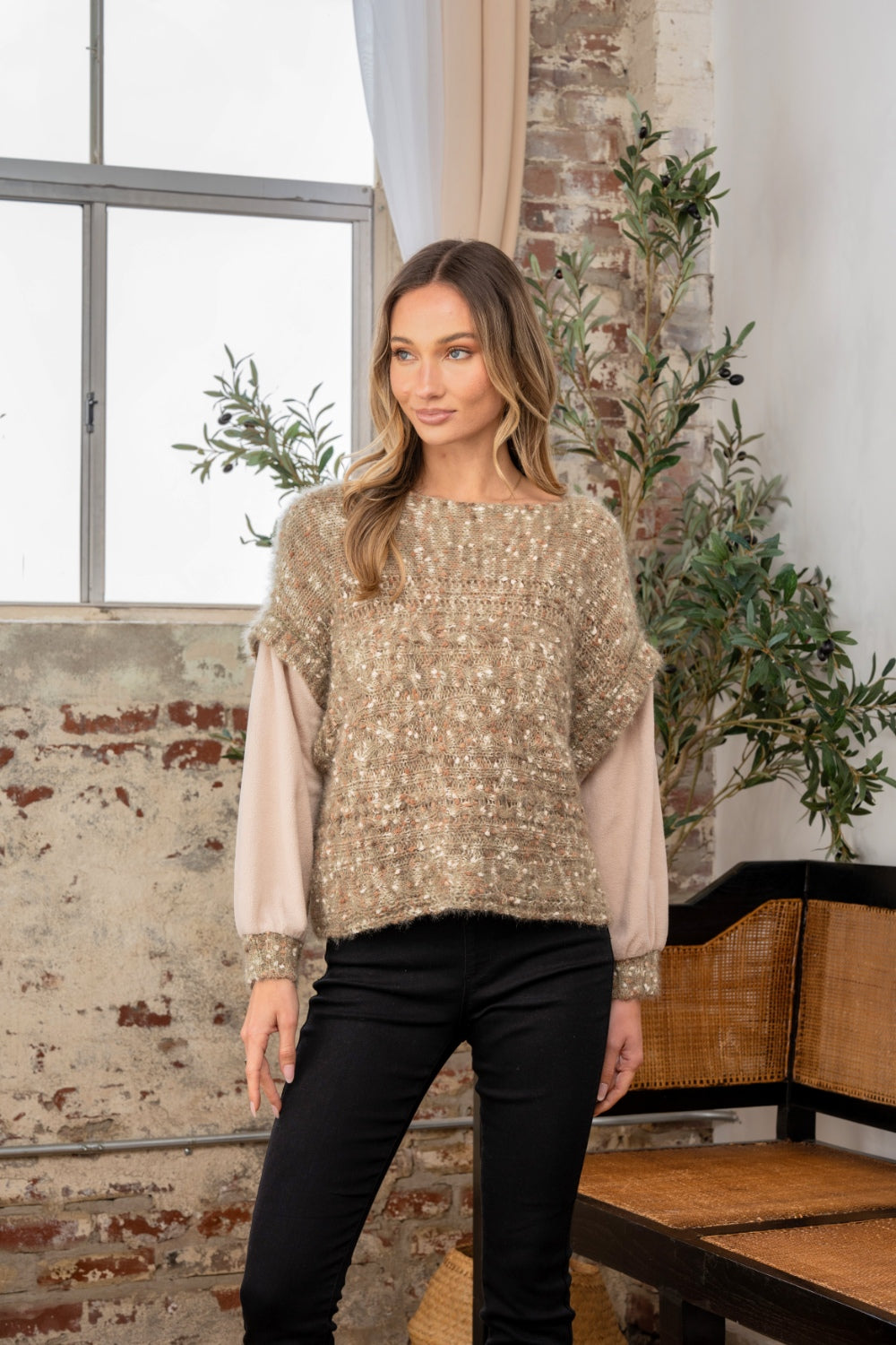 Standing indoors by a window, a person looks cozy and stylish in black pants, their textured sweater contrasting perfectly with the brick wall and greenery backdrop. The Sew In Love Full Size Fuzzy Long Sleeve Knit Top adds an elegant touch as one hand rests in their pocket.