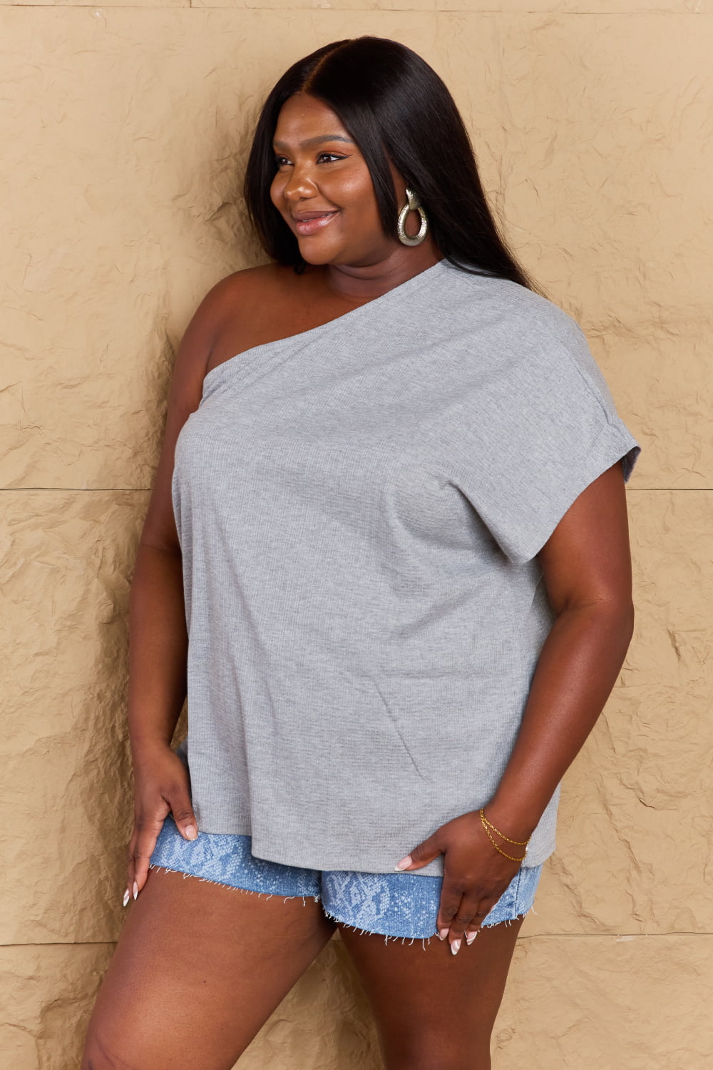 A person is posing against a beige textured wall, wearing the Ninexis in My Groove One Shoulder Loose Top made from lightweight gray fabric, denim shorts, and brown sandals. They have a hand on their hip and are smiling at the camera.