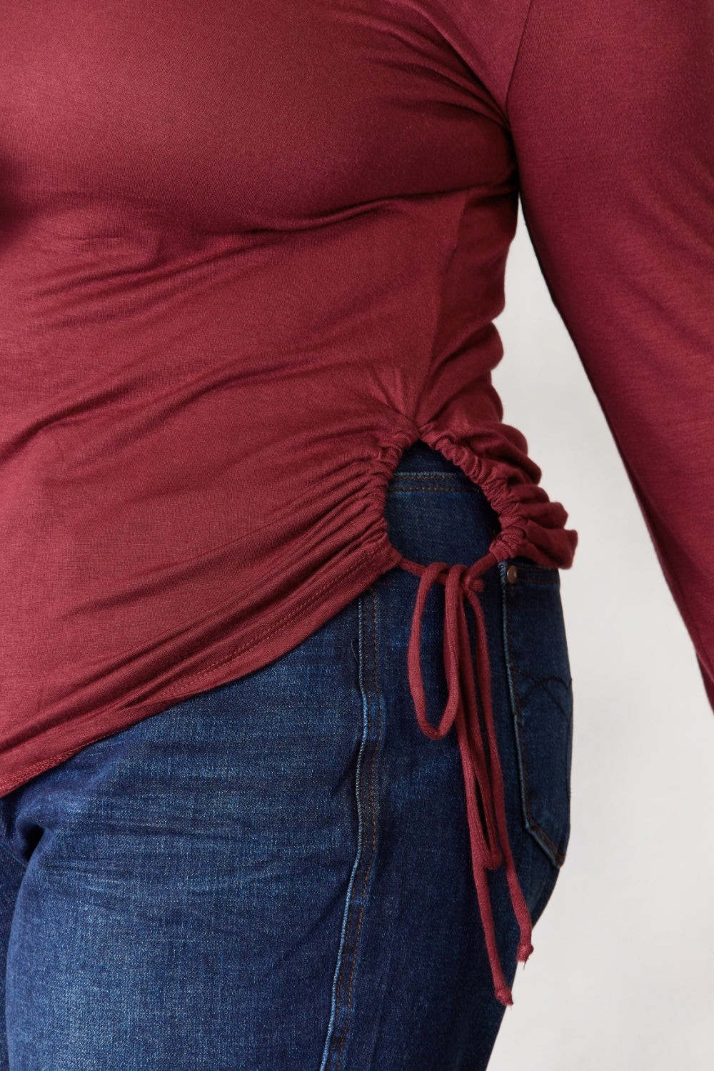 A woman stands in front of a plain background and a small tree, wearing the Culture Code Full Size Drawstring Round Neck Long Sleeve Top in maroon and blue jeans.