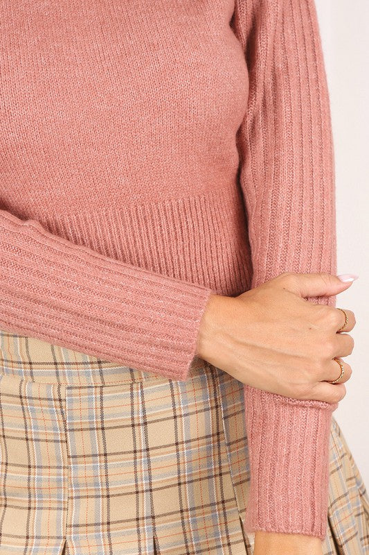 A person wearing a pink crop mock neck sweater with raglan sleeves poses with one hand near their chin.