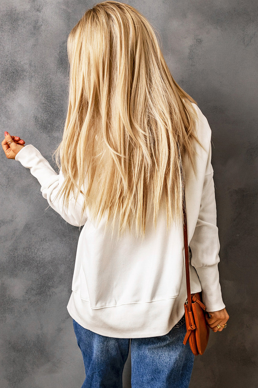 A person with long blonde hair, wearing a White Batwing Sleeve Pocketed Henley Hoodie and blue jeans, stands with their back to the camera, holding a small brown bag over their shoulder. The hoodie features a subtle patchwork design that adds an interesting texture to the outfit.