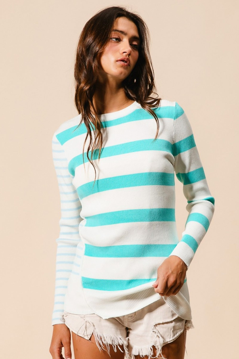 A person with long hair is wearing a BiBi Contrast Striped Asymmetrical Hem Knit Top featuring bold white and teal stripes, paired with white distressed shorts, and stands against a beige background.