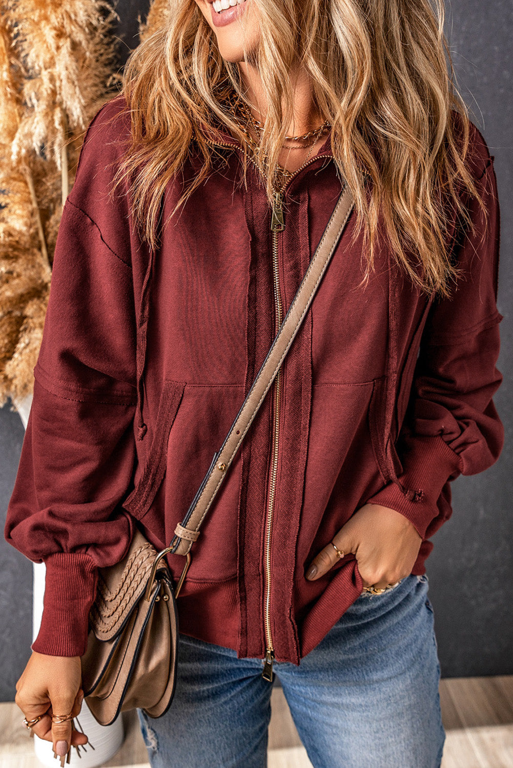 A woman with long, wavy blonde hair is seen from behind, wearing a trendy Brown Raw Edge Exposed Seam Full Zip Hoodie and carrying a brown shoulder bag.