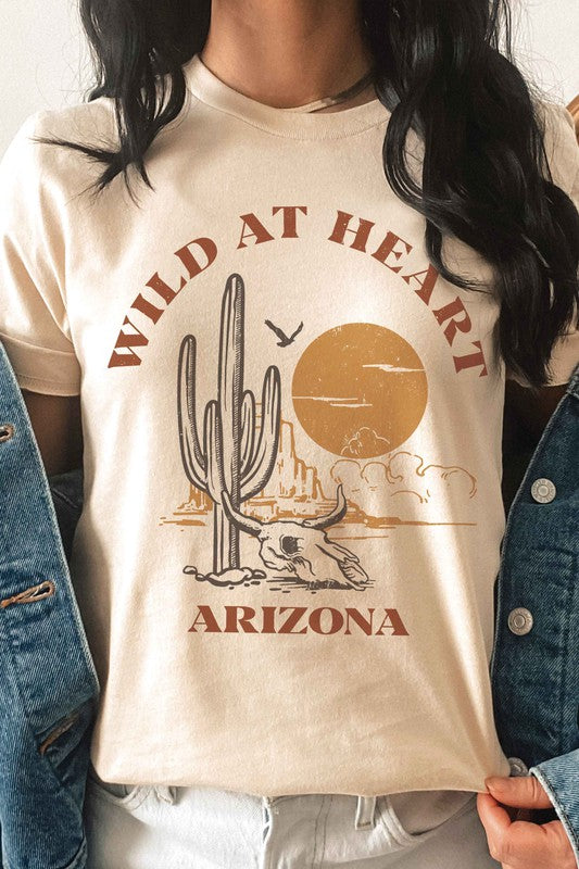 Woman wearing the WILD AT HEART ARIZONA Graphic Tee in white, which features the text "Wild at Heart Arizona" along with an illustration of a cactus, a sun, and a lounging skeleton. This tee is made from 100% cotton and offers unisex sizing for ultimate comfort and style.