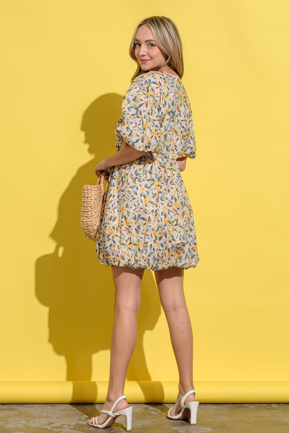 A person wearing the And The Why Full Size Floral Surplice Puff Sleeve Dress stands against a yellow background, holding a woven bag and wearing white sandals.