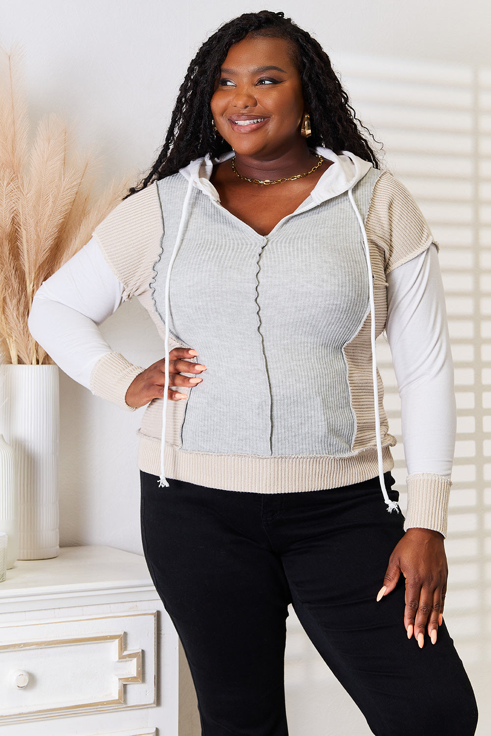 A woman wearing the Double Take Color Block Exposed Seam Drawstring Hoodie poses indoors. She has long braided hair, is smiling, and rests her hand on her hip.