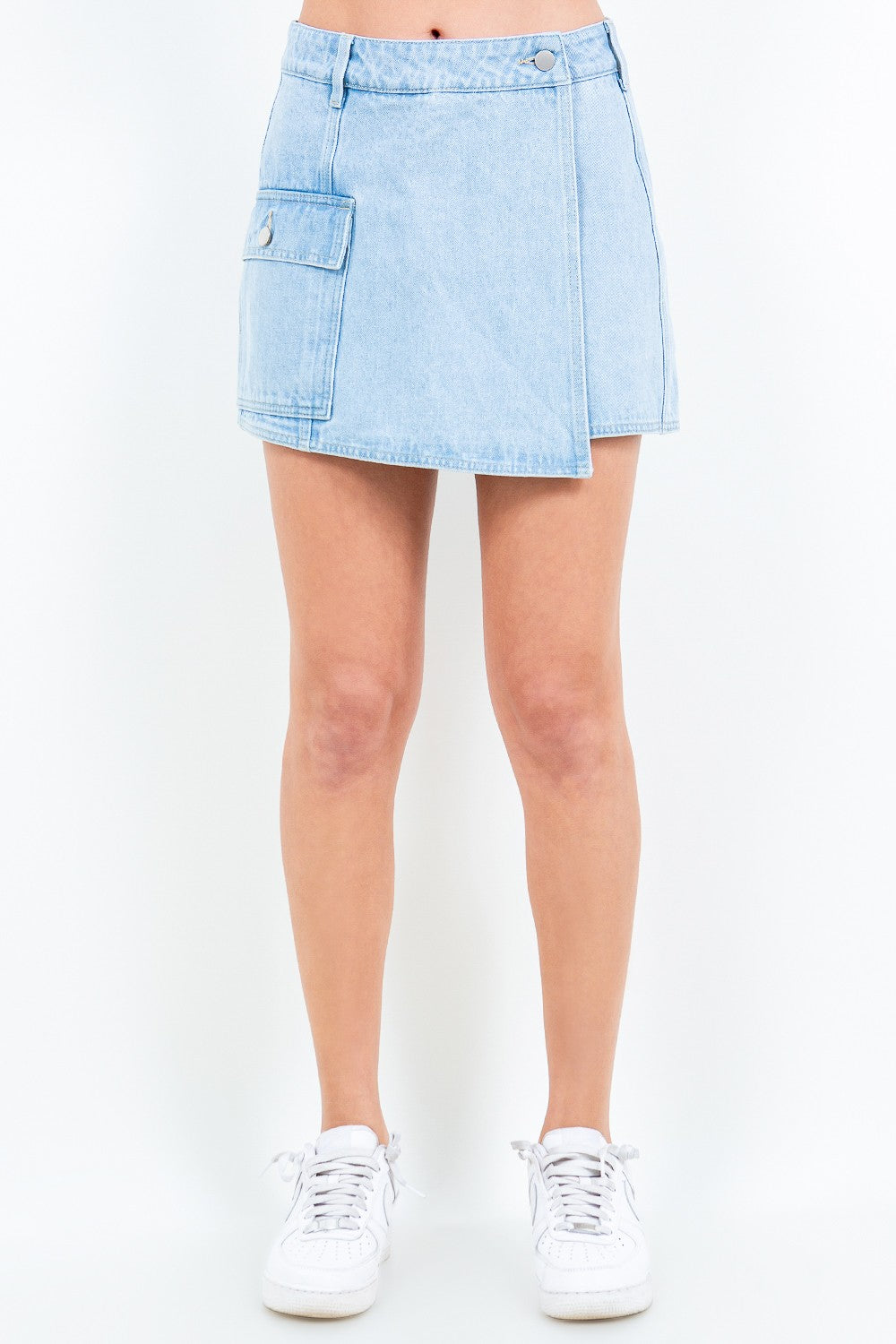 A person wears the American Bazi Cargo Pocket Mini Skort in light blue denim, featuring a large front pocket and button waist closure, paired with white sneakers.