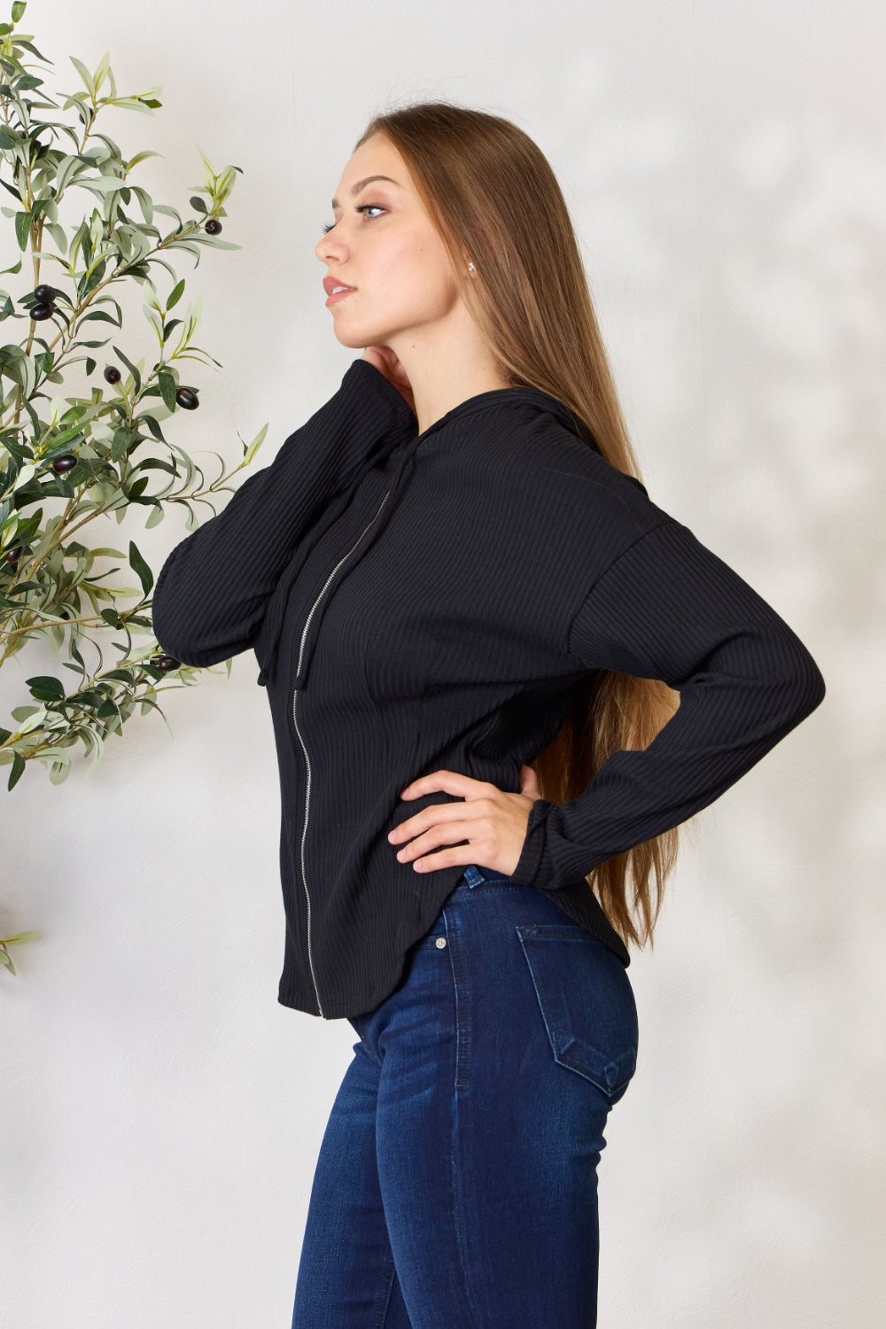 A person wearing the Culture Code Full Size Ribbed Zip Up Drawstring Hooded Jacket and blue jeans stands in front of a plain background with a small leafy plant to the left.