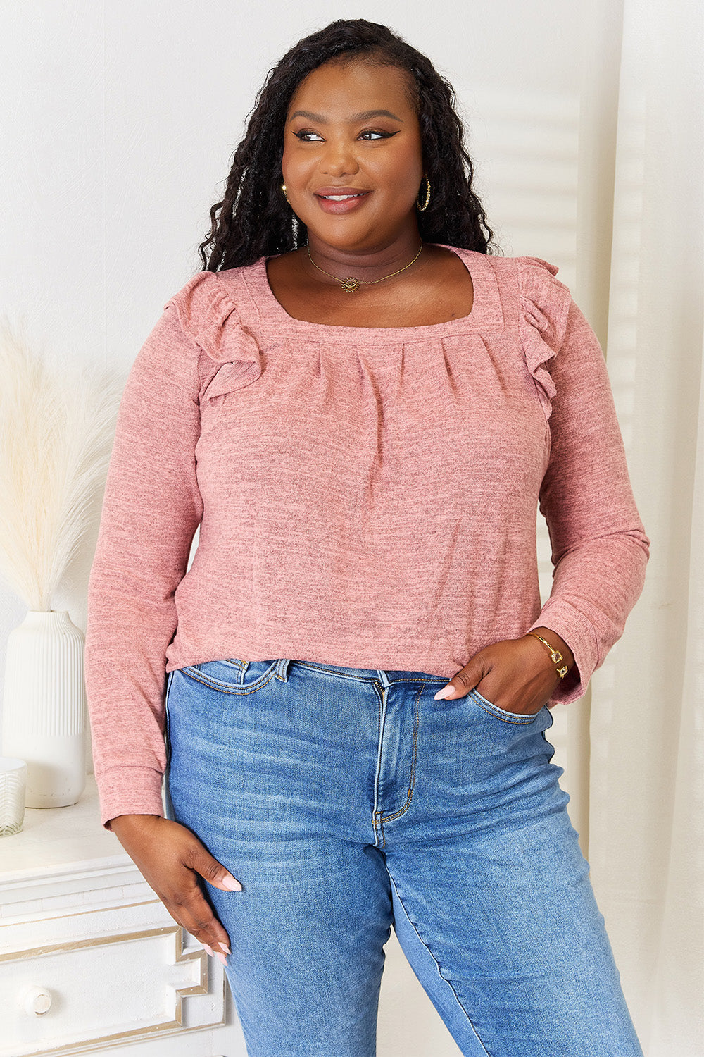 A woman wearing a Double Take Square Neck Ruffle Shoulder Long Sleeve T-Shirt in pink and blue jeans stands with one hand in her pocket, smiling. She has long curly hair and wears a necklace and bracelet. She stands in a room with white decor.