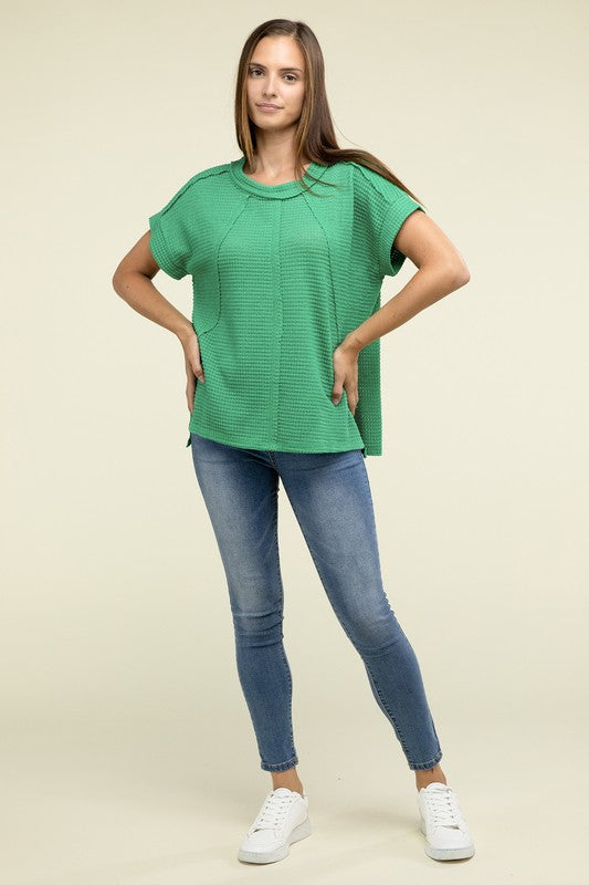 A woman with long brown hair is wearing a purple Brushed Waffle Exposed-Seam Short Sleeve Top and blue jeans, standing against a plain beige background. This casual wardrobe choice, with its short sleeves and side slits, is perfect for everyday wear.