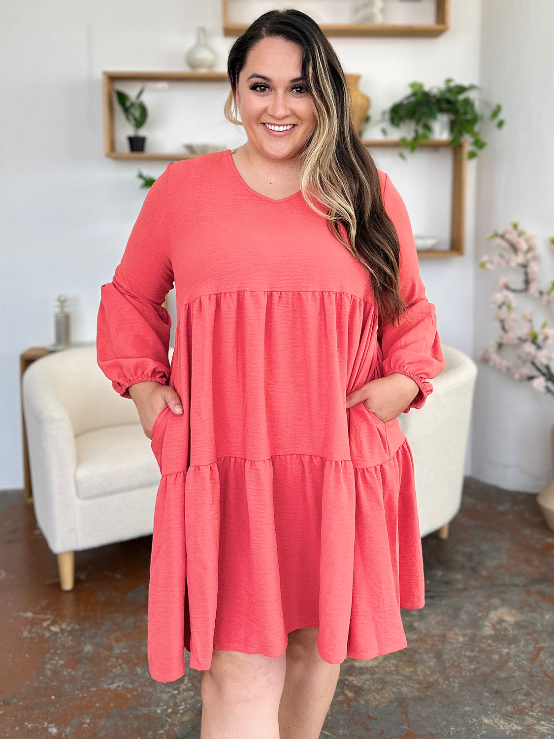 A woman stands wearing the Double Take Full Size V-Neck Balloon Sleeve Tiered Dress with Pockets in pink. Crafted from high stretch yarn, this dress combines comfort and style effortlessly. She has long, wavy hair and accessorizes with hoop earrings and a necklace. This imported garment is easy to care for – simply machine wash cold to keep it looking fresh.