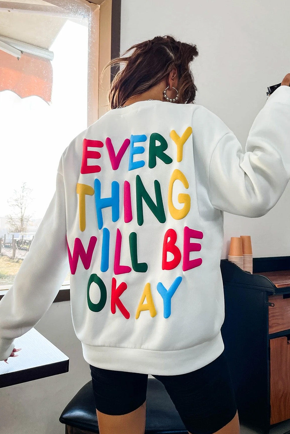 A person wearing a white beanie, the EVERY THING WILL BE OKAY Colorful Letters Sweatshirt, and sunglasses, holding a coffee cup, stands outside near windows.