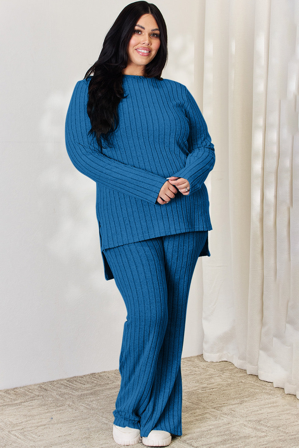 A person wearing the Basic Bae Full Size Ribbed High-Low Top and Wide Leg Pants Set in light gray, made from stretchy material, stands indoors in front of a white curtain.