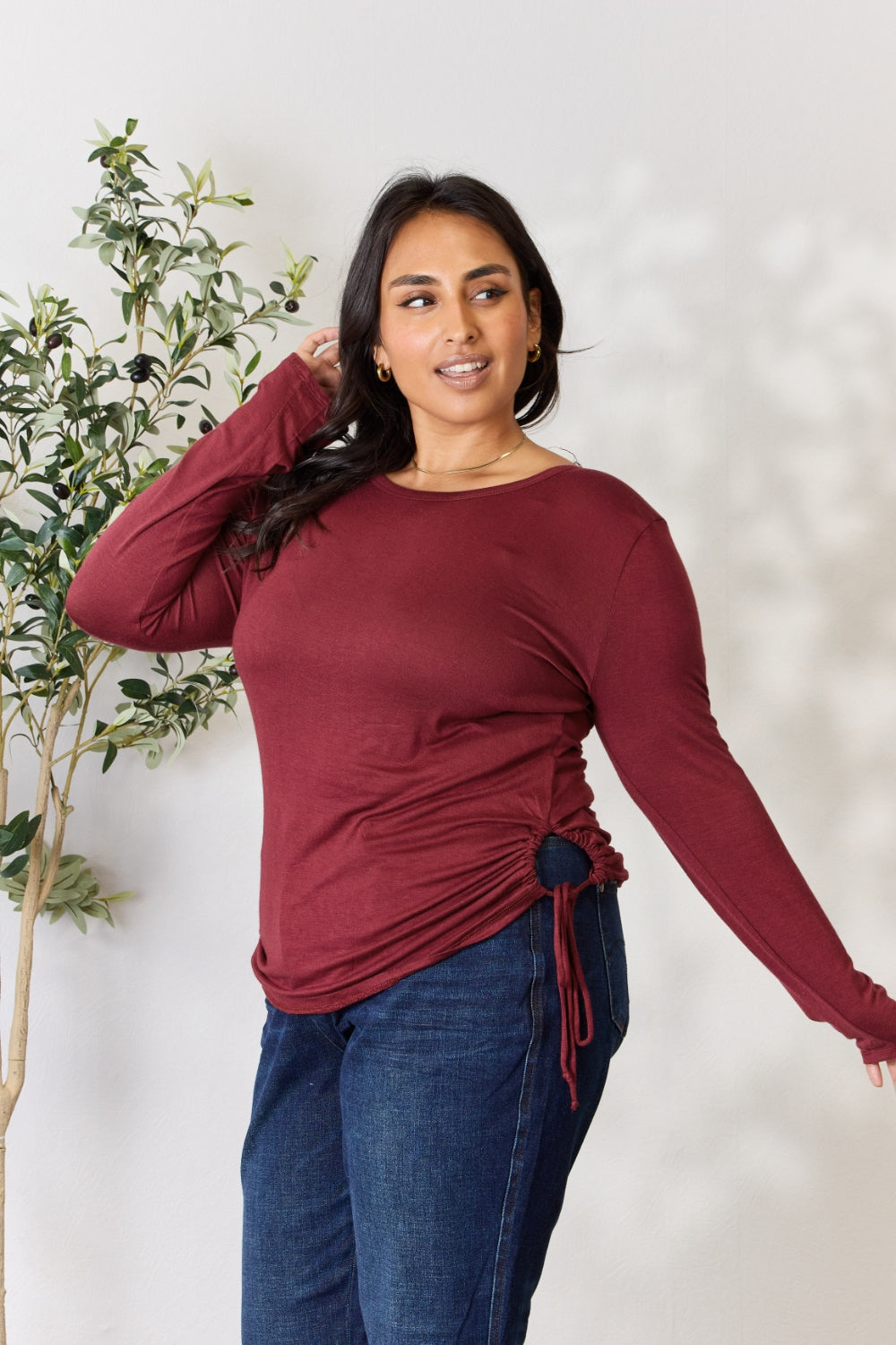 A woman stands in front of a plain background and a small tree, wearing the Culture Code Full Size Drawstring Round Neck Long Sleeve Top in maroon and blue jeans.