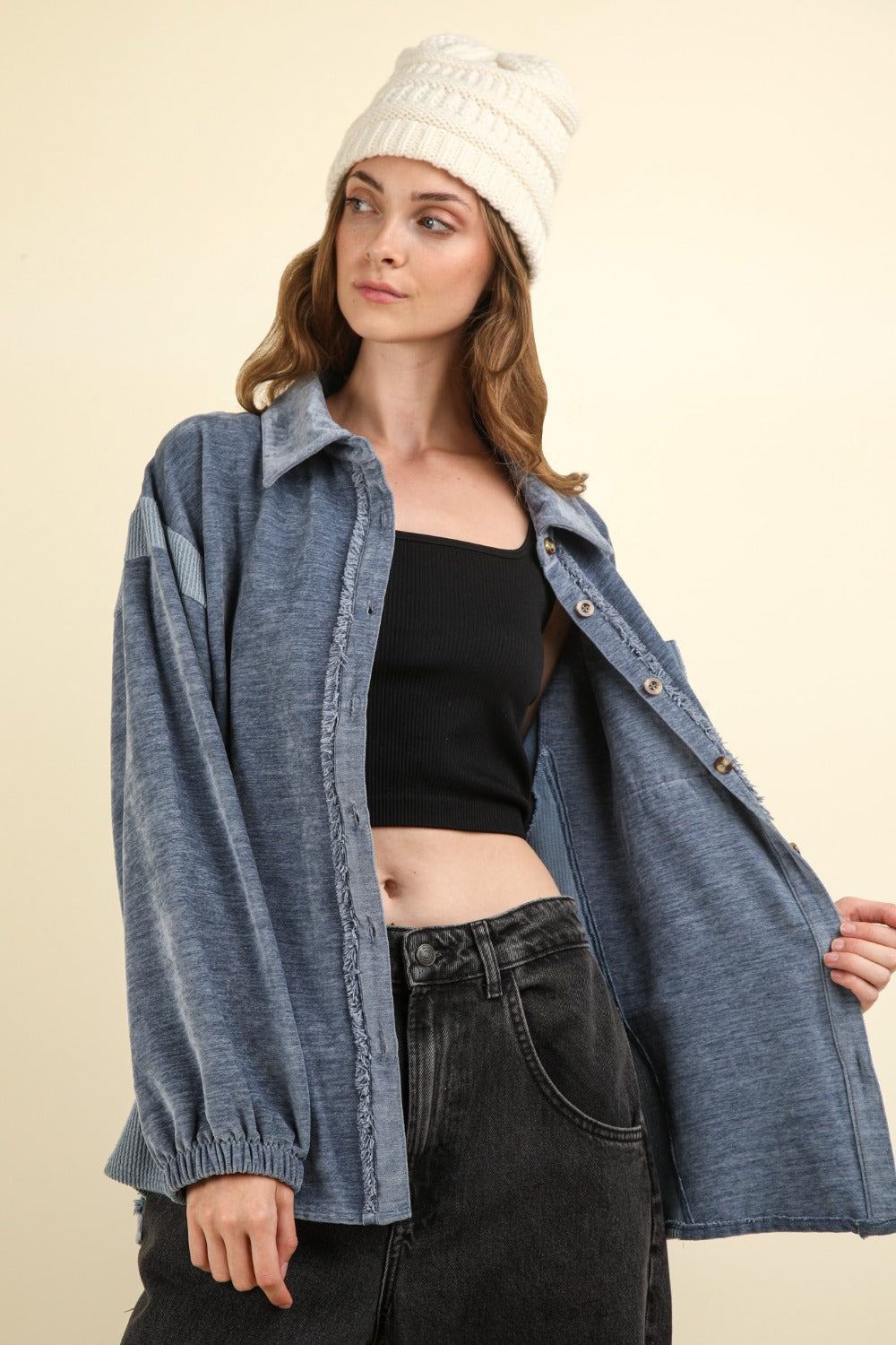 A person wearing the VERY J Mixed Media Button Down Raw Hem Shacket over a black crop top and black jeans, with a white knit hat. The background is plain and light-colored.