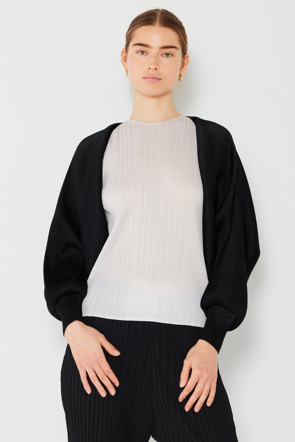 A person stands wearing the stylish Marina West Swim Rib Pleated Puff Sleeve Bolero Cardigan in beige, featuring draped sleeves, paired with a trendy black pleated top.