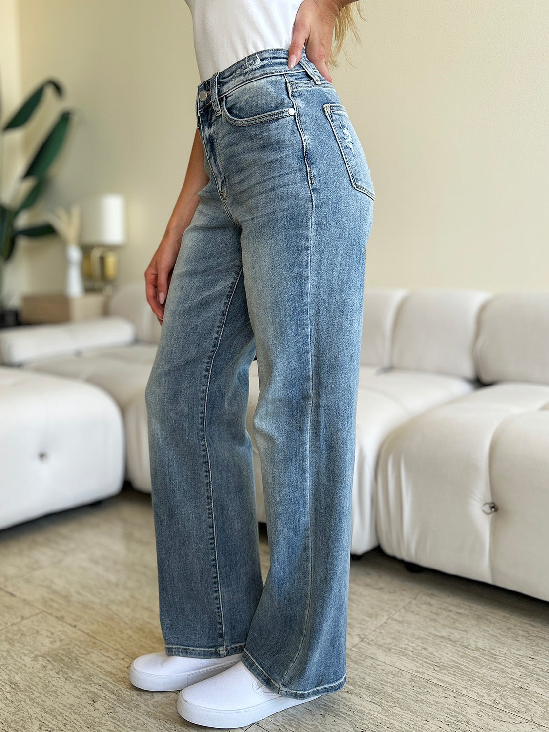 A person wearing Judy Blue Full Size High Waist Straight Jeans and white shoes is standing indoors. There is a white couch and a large plant in the background.