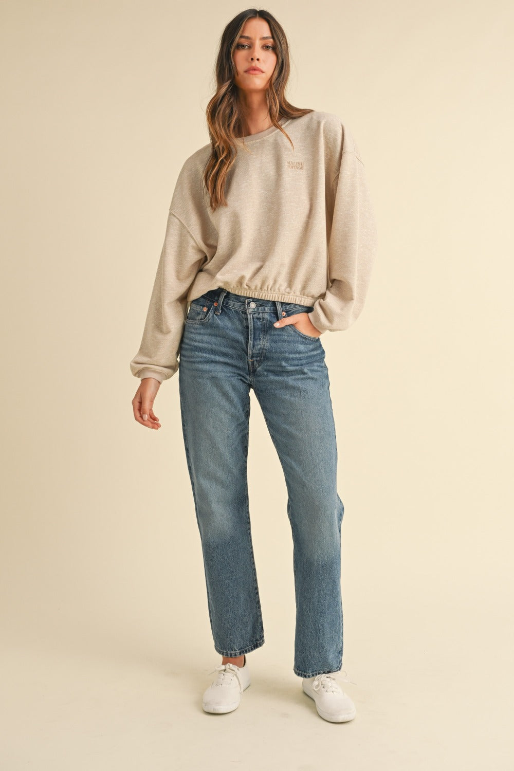 A woman with long brown hair wears the Mable Round Neck Letter Embroidery Crop Sweatshirt in beige, paired with blue jeans, standing confidently with one hand in her pocket against a plain background, perfectly capturing effortless street style.