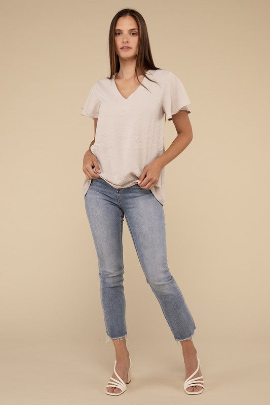 A woman with long straight hair models the Woven Airflow Flutter Sleeve Top in green, matched with light blue jeans, as she poses with her hands in her pockets against a plain beige background.