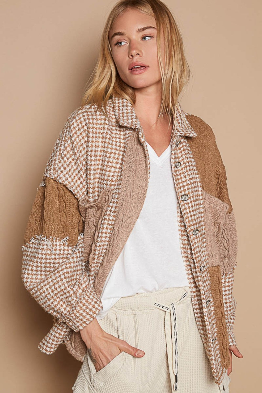 A woman with long blonde hair wears the POL Houndstooth Contrast Cable Knit Pocket Shacket, featuring a patchwork design in various shades of brown and beige, over a white V-neck shirt and paired with light-colored pants. She is standing against a neutral background.