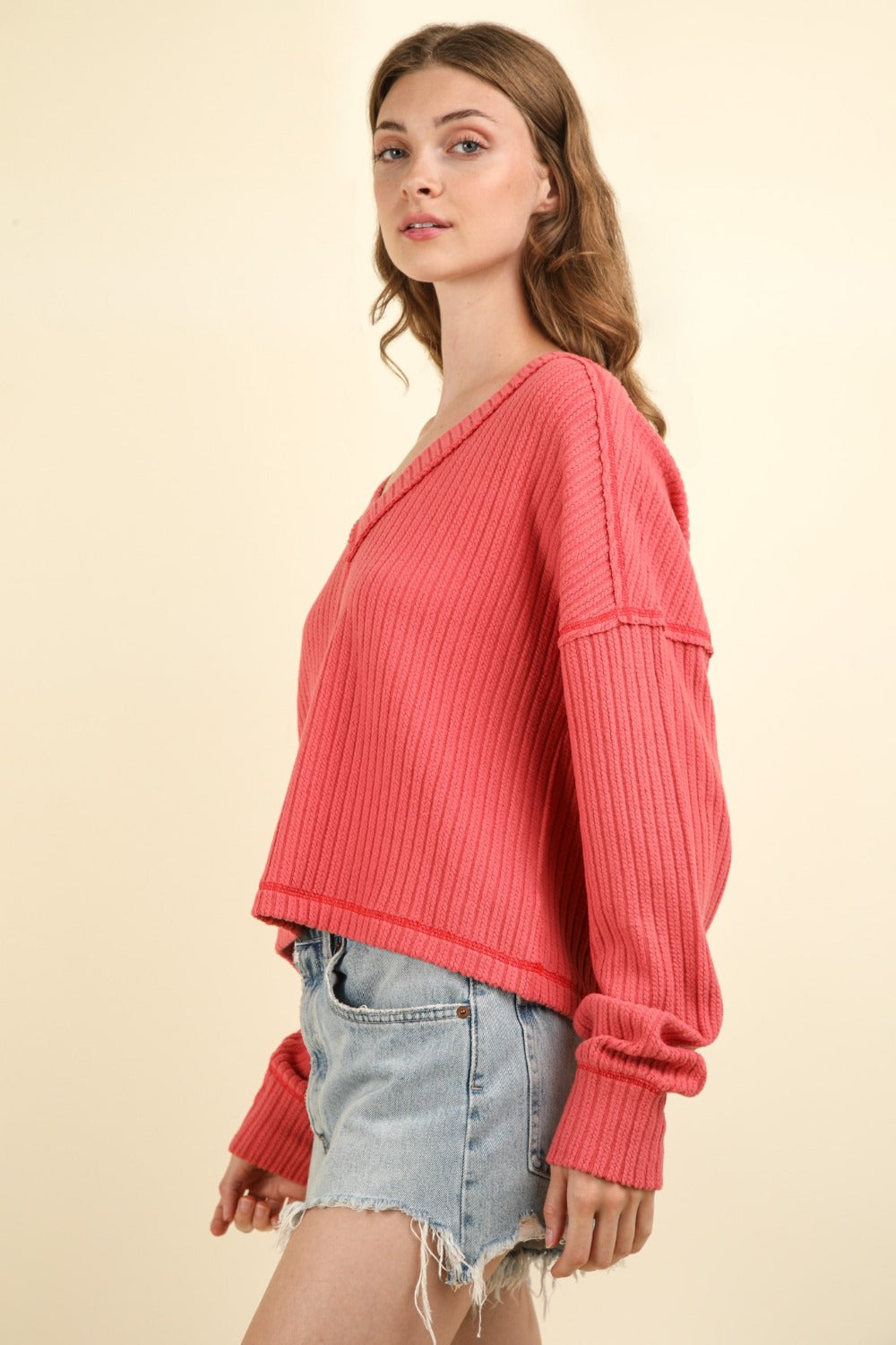 A person with long hair wearing a pink VERY J Exposed Seam V-Neck Ribbed Knit Top and blue jeans stands against a plain background.