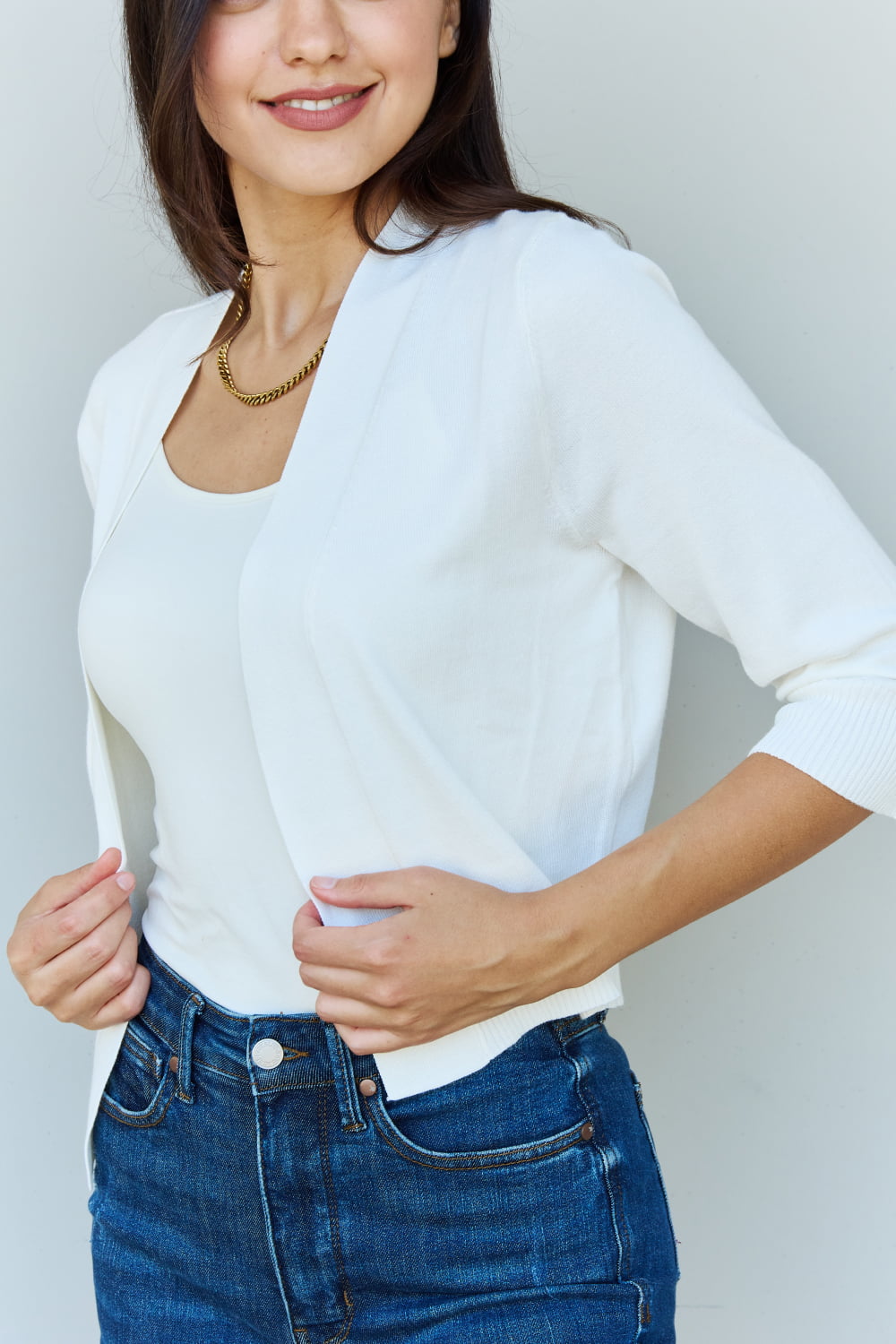 A stylish person wearing sunglasses, a Doublju My Favorite Full Size 3/4 Sleeve Cropped Cardigan in Ivory, and blue jeans poses in front of a plain background, effortlessly highlighting their versatile look.