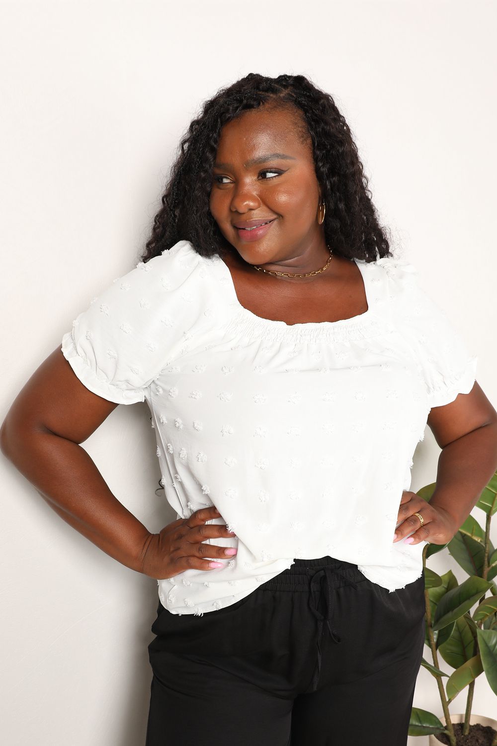 A person wearing the Mandy Swiss Dot Puff Sleeve Square Neck Blouse in white, paired with black pants, stands in front of a light background with green leaves visible on the right side.