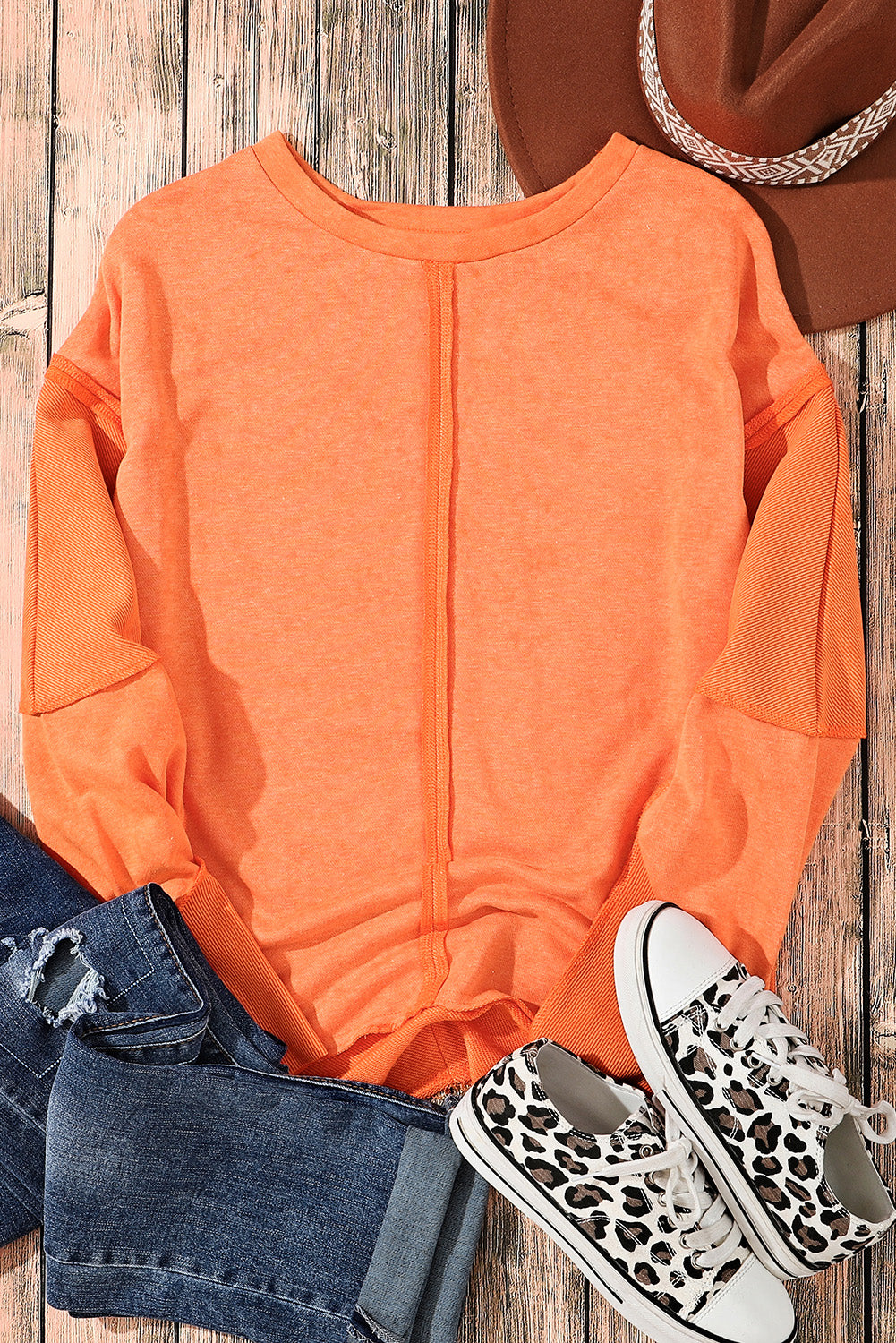 A person with long blonde hair is standing with their back to the camera, wearing an Orange Exposed Seamed High Low Raw Edge Sweatshirt and denim shorts, in front of a brick wall.