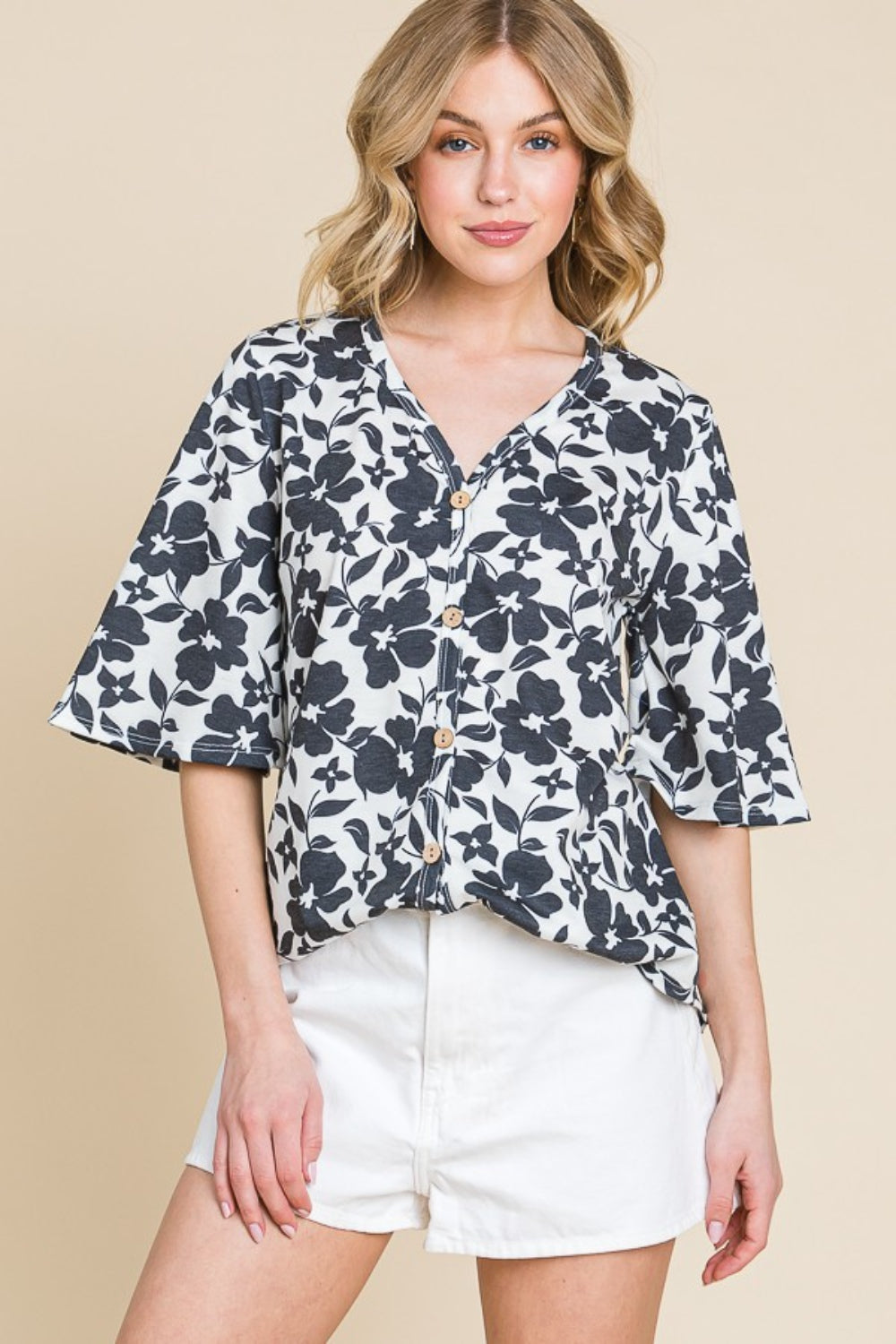 A person stands against a beige background, wearing the BOMBOM Floral Decorative Button V-Neck Top paired with white shorts, looking at the camera.