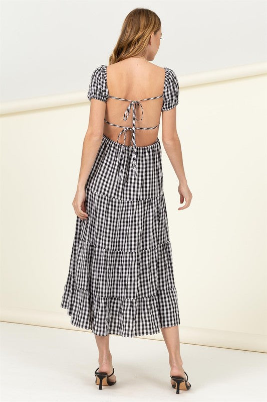 Wearing the "Somewhere to Go Tie-Back Gingham Print Maxi Dress," which features a black and white gingham print with puffed sleeves, a tiered skirt, and elegant tie-back detail, a person poses stylishly paired with black sandals.