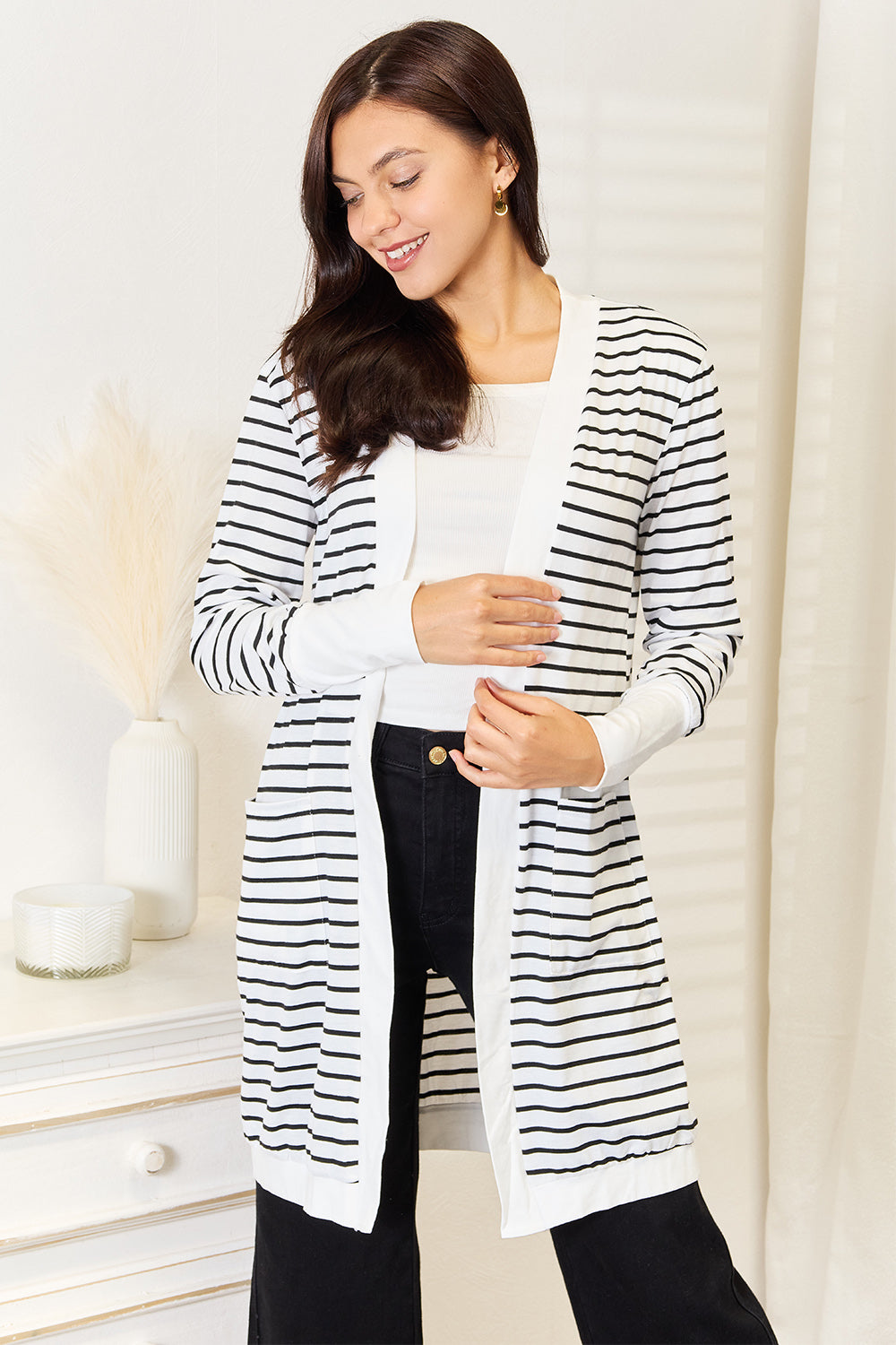 A woman wearing the Double Take Striped Open Front Longline Cardigan over a white top and black pants stands indoors, smiling at the camera. She is posing with one hand on her hip, showcasing this versatile piece.